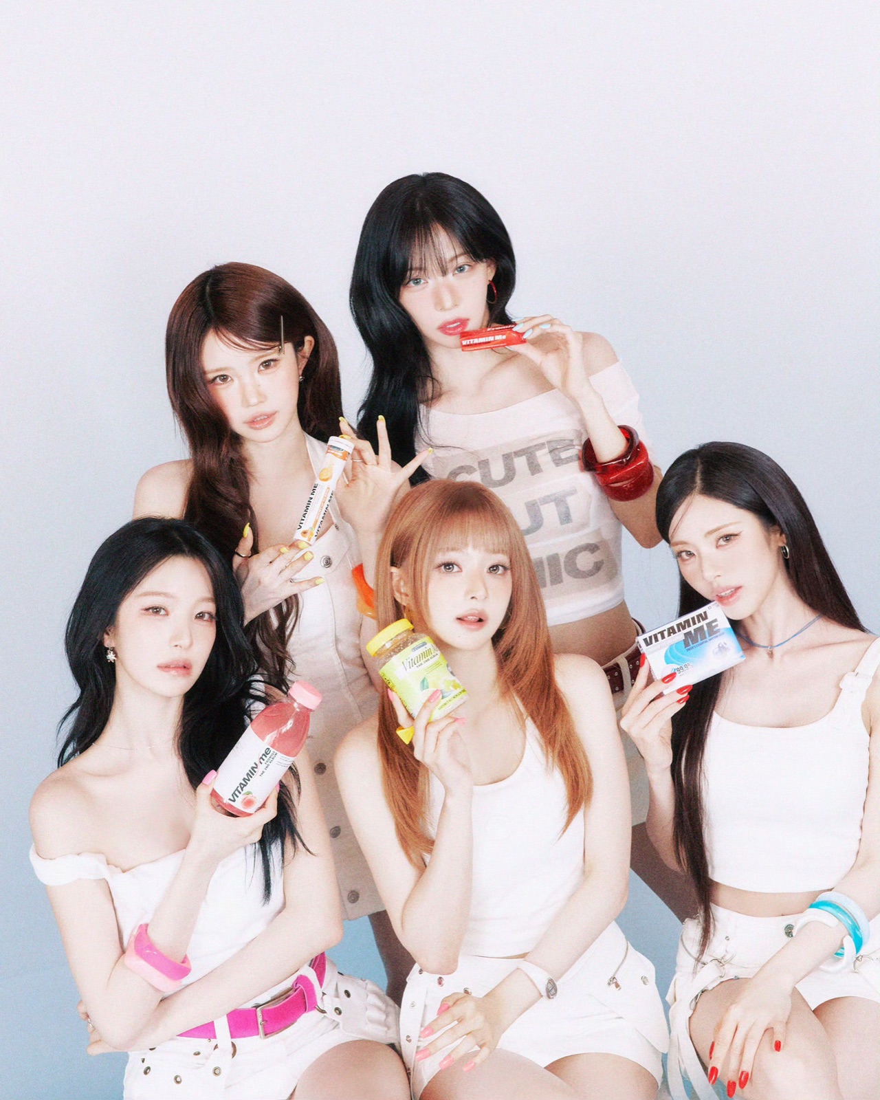 Five fromis_9 members dressed in white and holding colourful Vitamin Me packages.