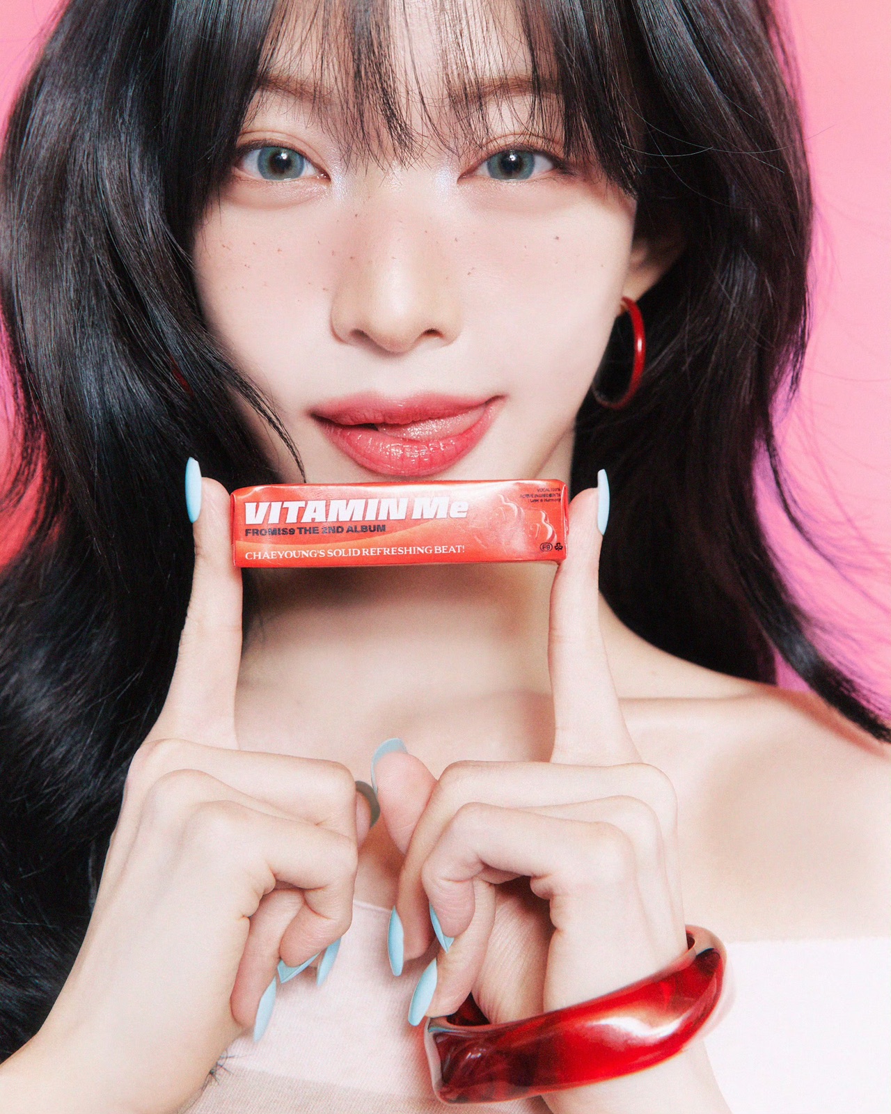 Close portrait of a fromis_9 member holding a red Vitamin Me package against a pink background.