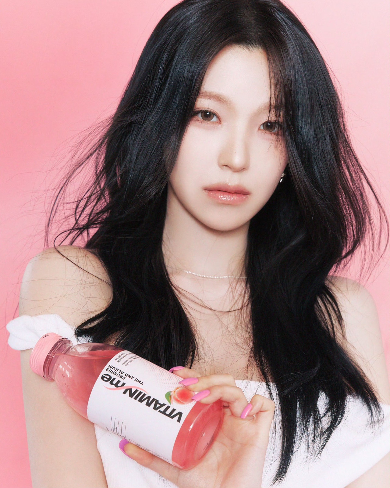 A fromis_9 member holding a pink Vitamin Me bottle against a pink background.