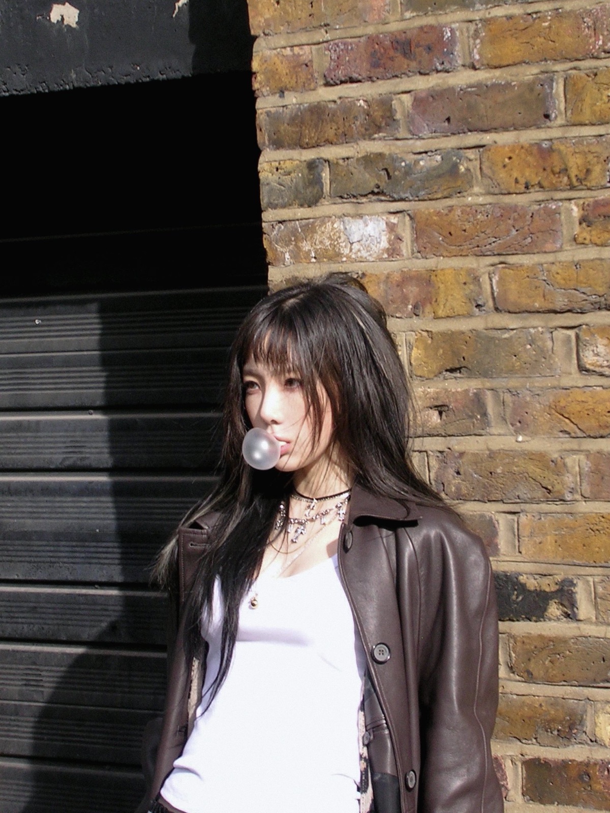 Taeyeon blowing a bubble beside a sunlit brick wall and dark doorway.